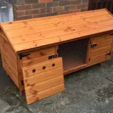 The 5ft British Giant Large Rabbit Hutch, designed with a fox-proof wooden structure, slanted roof, mesh window, and open side door, is positioned on a concrete path adjacent to a brick wall.