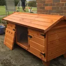 A 5ft British Giant Large Rabbit Hutch, designed to be fox-proof with an open door, is situated on a concrete patio next to a brick wall. In the blurred background, a person watches over near a grassy area and garden pots, making sure the rabbits remain safe and sound.