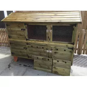 The 6ft British Giant XL Double Rabbit Hutch, featuring wire doors and complete winter protection, is designed with multiple compartments and mesh windows. It rests on a concrete surface near a wooden fence and could easily function as both a chicken coop and a rabbit hutch.