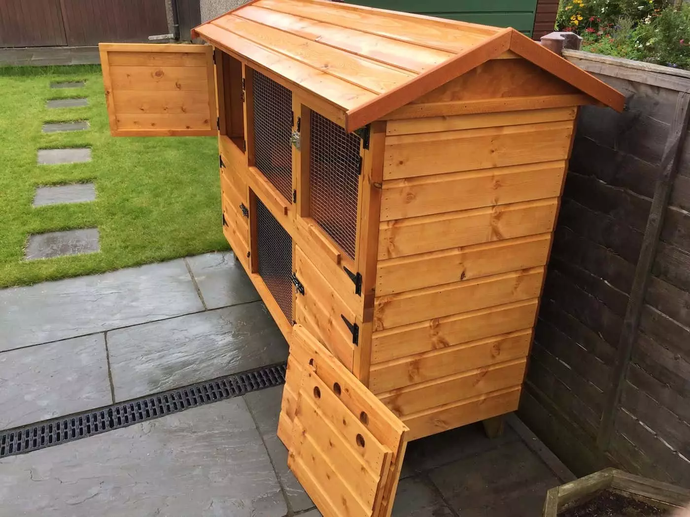 The 5ft British Giant Large Double Rabbit Hutch + Upstairs Winter Protection offers ample space with its wooden design, featuring multiple compartments and open doors with mesh panels. It's ideal for large breed rabbits and can be perfectly placed on a patio adjacent to a grass lawn.