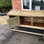 Wooden chicken coop with open doors on a paved patio area, adjacent to a brick wall and window, seamlessly integrating into a cozy space ideal for winter rabbit care.